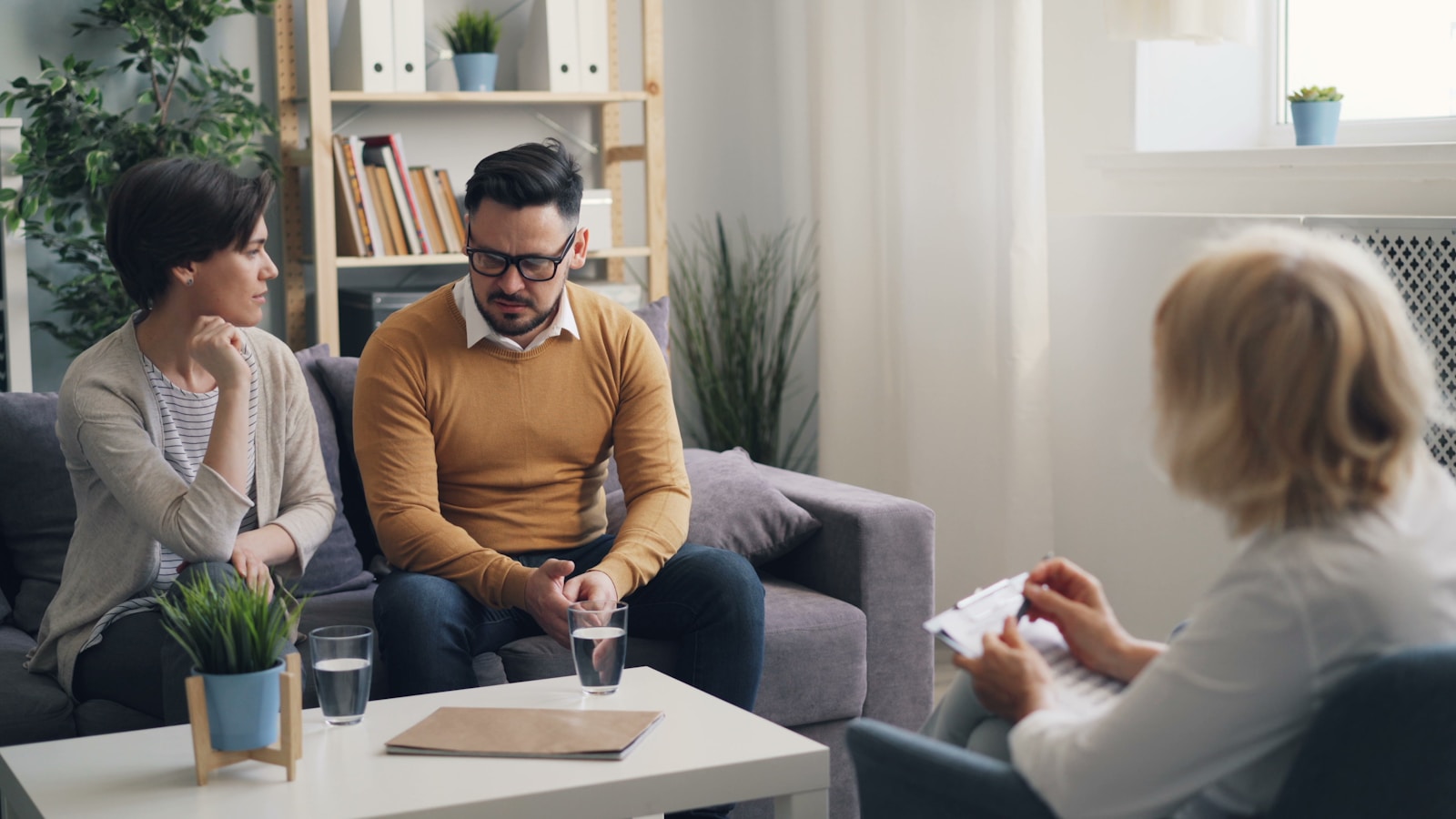 A couple sits on a couch talking with an older counselor in a warm, book-lined office.